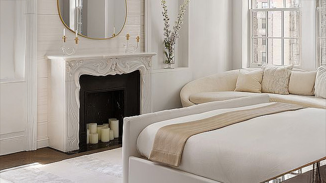 This is a bright and elegant primary bedroom featuring a fireplace with a white mantle and a decorative mirror. There's a curved sofa nestled in the corner by a window, maximizing the room's comfort and style. The bed is neatly made with neutral linens, reinforcing the room's inviting atmosphere.