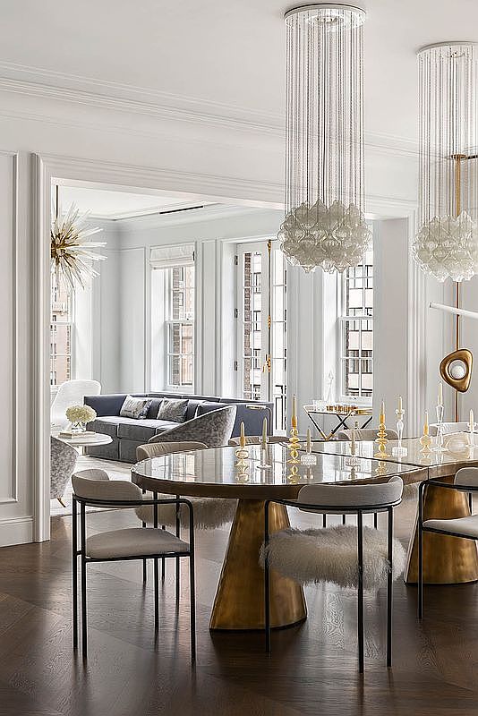 This elegant dining room features a large, round table with a reflective surface and a unique, sculptural base, surrounded by modern chairs with sleek black frames and light-colored upholstery. Above the table hang two striking chandeliers with cascading crystal strands, adding a touch of glamour. The room opens into a living area, creating a seamless and sophisticated living space.