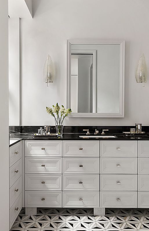 This is a well-lit primary bathroom featuring a double vanity with white cabinetry and a black countertop. A large mirror hangs above the sinks, flanked by decorative sconces. The floor is tiled with a geometric pattern, adding a touch of elegance to the space.