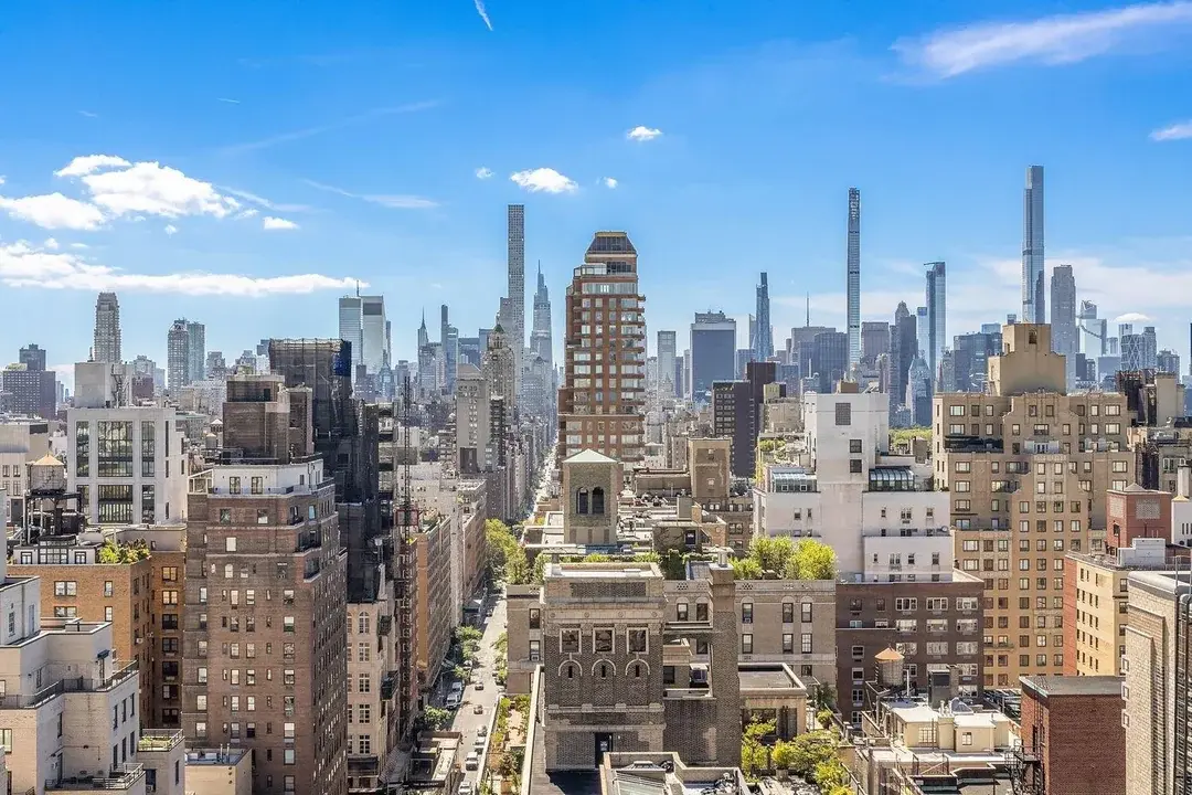 The image showcases an expansive aerial cityscape, dominated by an array of towering skyscrapers under a clear blue sky with scattered clouds. The foreground features dense urban buildings, offering a sense of the scale and density of the city. This view emphasizes the location's proximity to the city center and potential for stunning views.