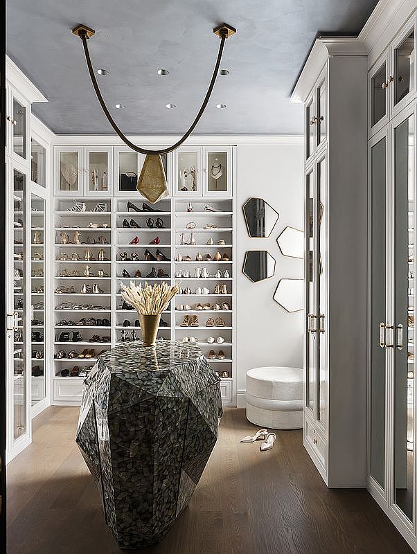 This is a luxurious walk-in closet featuring custom white cabinetry with glass doors, providing ample storage for shoes and accessories. A unique, faceted center island adds a touch of modern elegance, while a statement pendant light fixture hangs above. The overall impression is one of high-end design and organization.