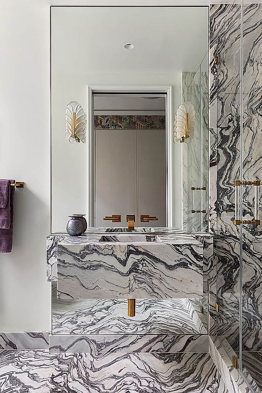 This is a luxurious primary bathroom featuring striking marble walls, flooring, and vanity. The vanity is integrated with a large mirror and flanked by elegant sconces. The design exudes a high-end, sophisticated aesthetic, perfect for a discerning buyer.