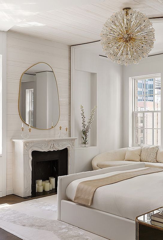 This is a bright and elegant primary bedroom featuring a large bed with white linens and a tan throw, a decorative fireplace with candles, and a unique gold-framed mirror. A crystal chandelier hangs from the ceiling, adding a touch of luxury. The room is bathed in natural light from a nearby window, creating a serene and inviting atmosphere.