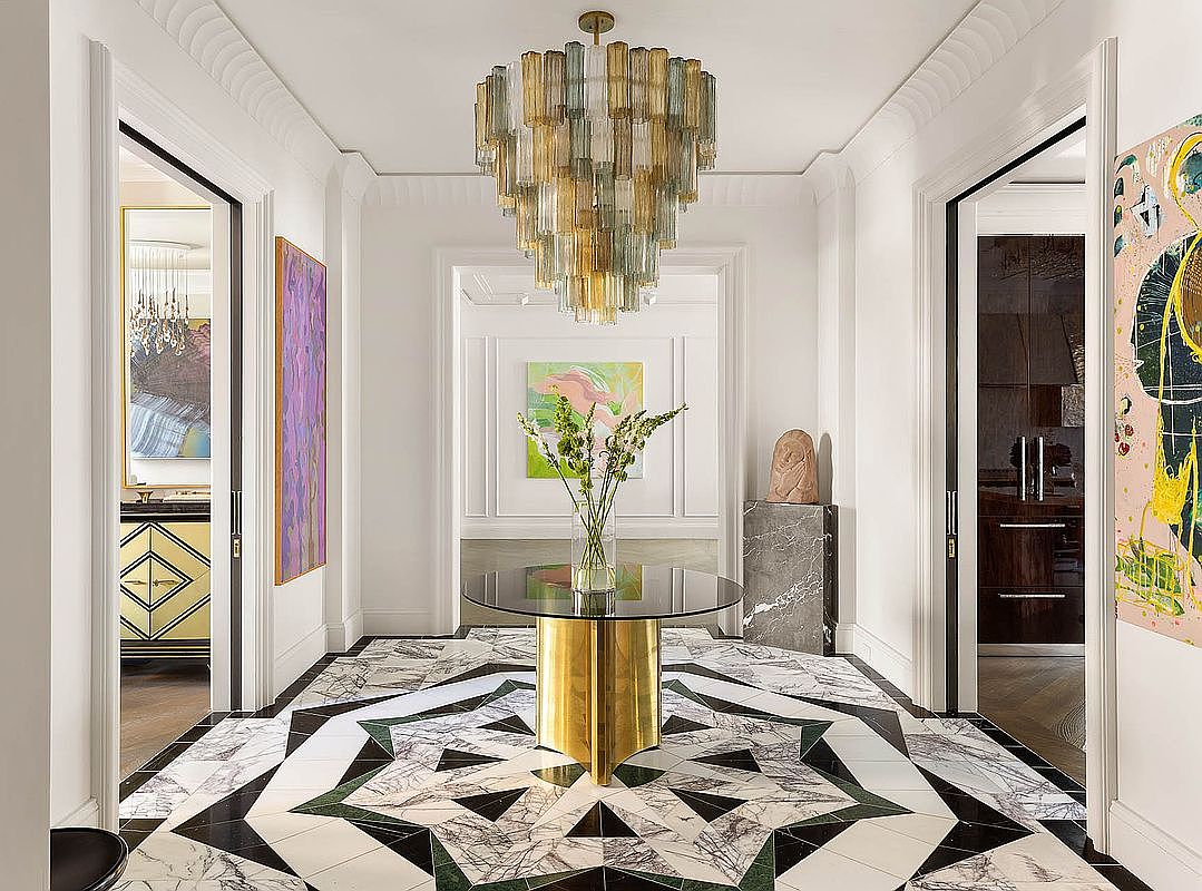 This is an interior shot of a luxurious hallway featuring a striking geometric patterned marble floor in black, white, and green. A modern chandelier hangs from the ceiling, illuminating the space, and artwork adorns the walls. A round glass table with a gold base sits in the center, adding to the elegant and sophisticated ambiance.