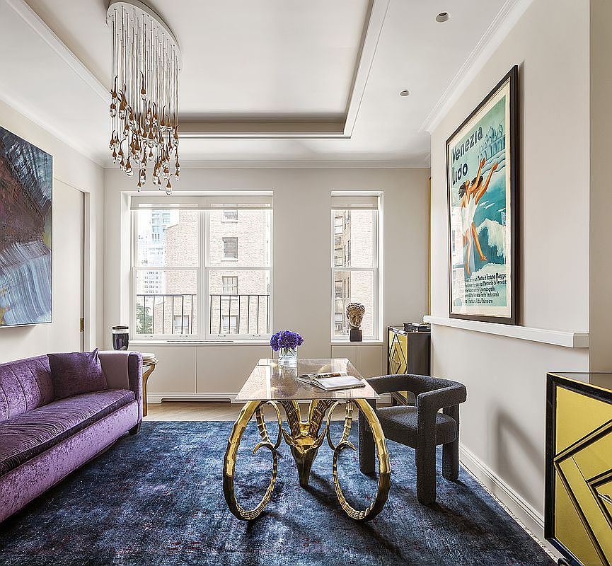 This interior shot showcases a stylish office or study area, featuring a purple velvet sofa, a unique gold-legged desk, and a modern gray chair. A striking chandelier hangs above, and a framed vintage poster adorns the wall, adding character to the space. The room is grounded by a deep blue rug, creating a sophisticated and inviting atmosphere.
