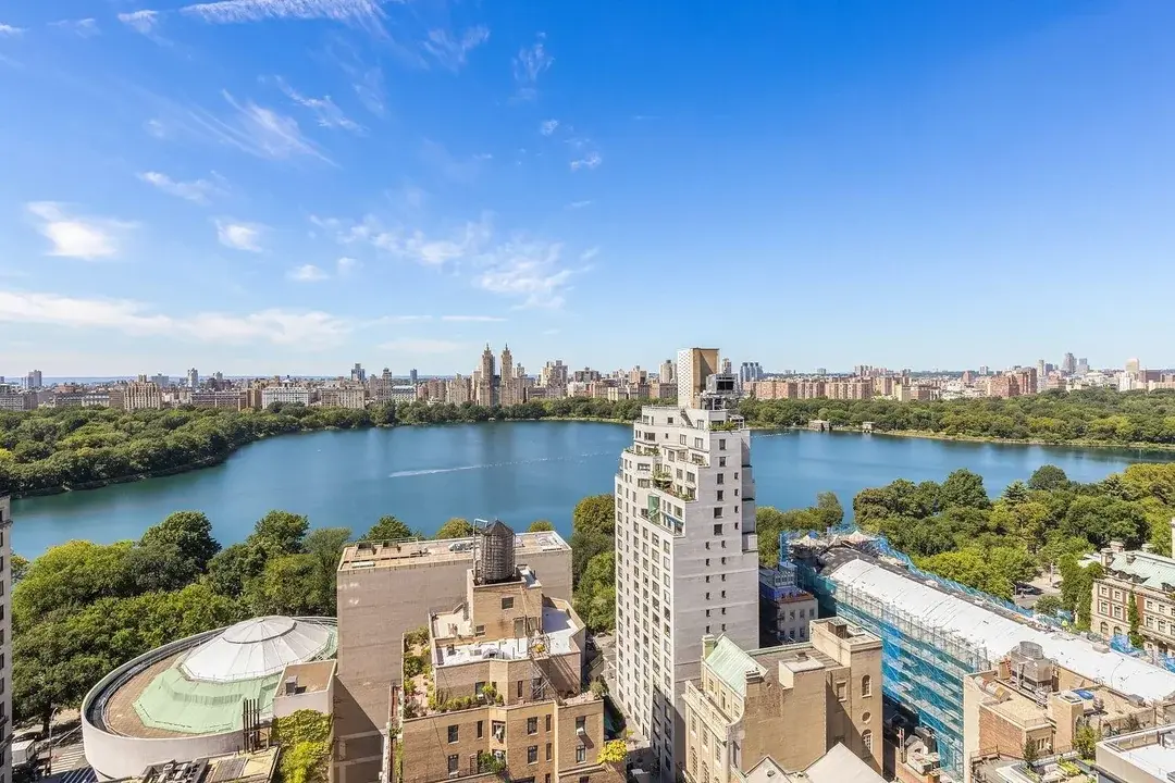 This aerial view showcases a stunning panorama of a city skyline, likely New York City, with a large lake and lush greenery in the foreground. Several high-rise buildings are visible, suggesting an urban environment with residential or commercial real estate. The image provides a sense of location, highlighting proximity to water and green spaces as desirable features.