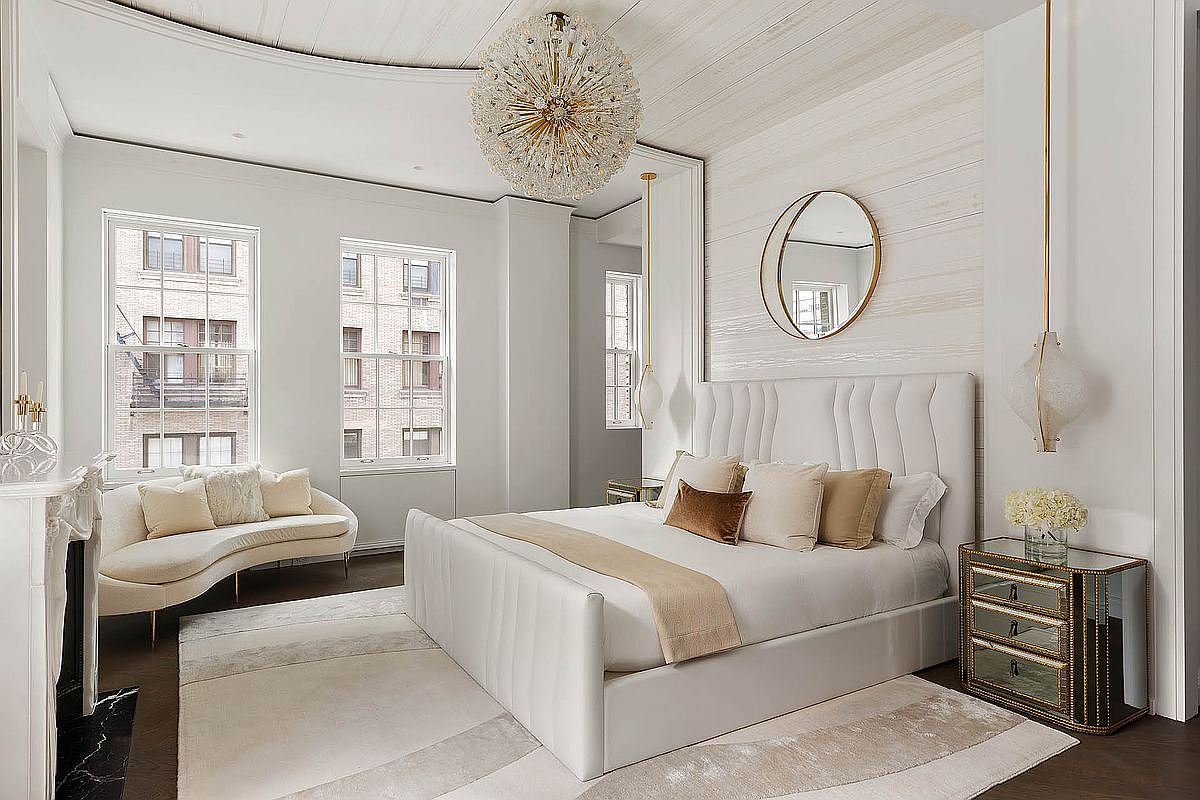 This is a luxurious primary bedroom featuring a large white upholstered bed with decorative pillows and a throw. The room is well-lit with natural light from the windows and features a unique chandelier and a round mirror above the bed. A curved sofa sits near the windows, and a mirrored nightstand is placed next to the bed, adding to the room's elegant and sophisticated ambiance.