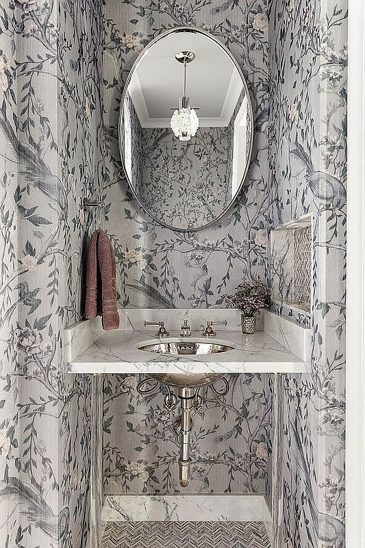 This is a stylish powder room featuring a marble countertop and sink with elegant fixtures. The walls are adorned with a sophisticated floral wallpaper, complemented by an oval mirror and a decorative light fixture. The overall impression is one of refined taste and attention to detail, making it a charming and inviting space.