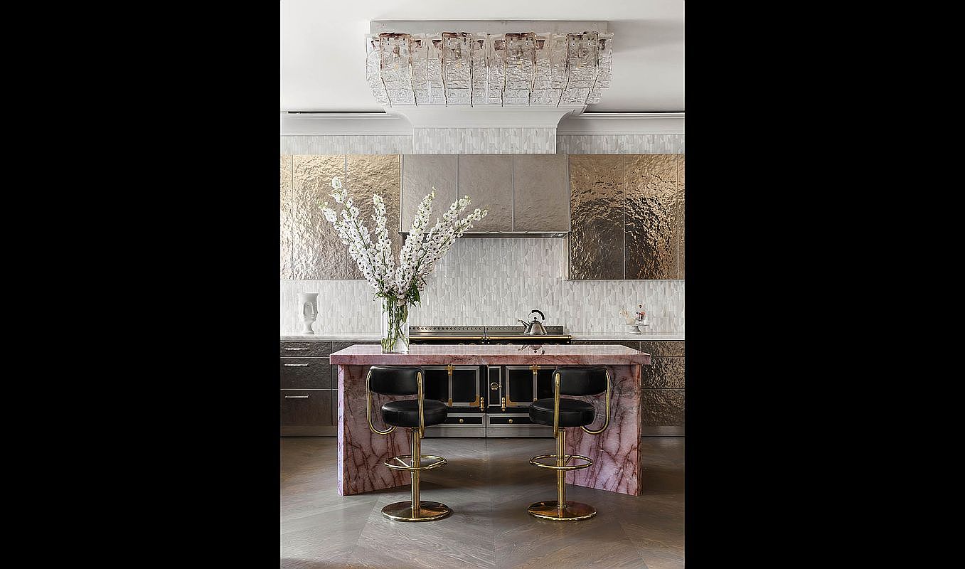 This is a stylish kitchen featuring a pink marble island with gold accents and black bar stools. The backsplash is a textured tile, and the cabinets are a metallic finish. A modern chandelier hangs above, and a large vase of white flowers sits on the island, adding a touch of elegance.