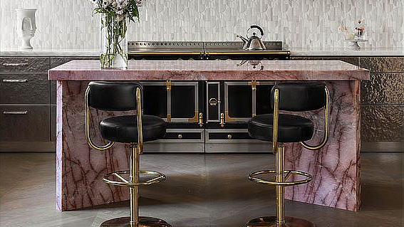 This image showcases a modern kitchen with a unique aesthetic. The centerpiece is likely a kitchen island with a pink marble countertop and matching base. Two black leather and gold bar stools are placed in front of the island, suggesting a seating area for dining or entertaining.