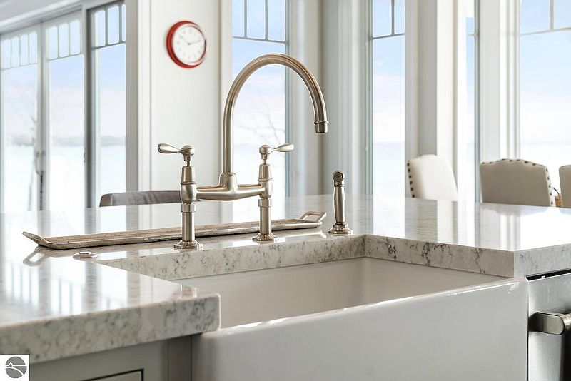 This image showcases a bright and modern kitchen featuring a farmhouse-style sink with a brushed nickel faucet. The countertops are a light-colored stone, and large windows provide ample natural light. The overall impression is clean, elegant, and inviting.