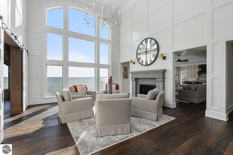 This is a bright and airy living room with a stunning view of the water through large windows. The room features a high ceiling with decorative paneling, a fireplace with a stone mantel, and comfortable seating arranged around a rug. The dark hardwood floors add warmth and contrast to the otherwise light and neutral color palette.