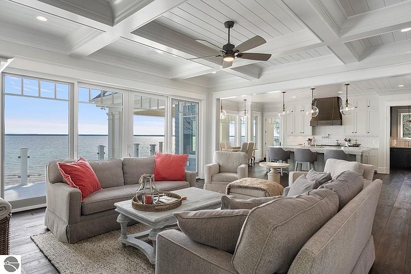 This is a bright and airy living room with a view of the water. The room features comfortable seating, a ceiling fan, and a neutral color palette. The open floor plan connects the living room to the kitchen, creating a spacious and inviting atmosphere.