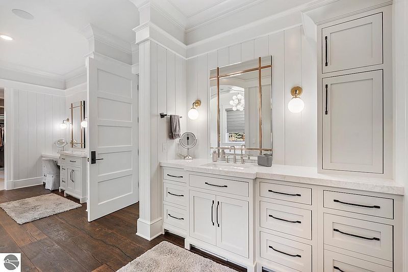 This is a well-lit primary bathroom featuring white cabinetry, marble countertops, and modern fixtures. The space includes a vanity area with a mirror and sconces, as well as built-in storage. The overall design is elegant and clean, creating a spa-like atmosphere.