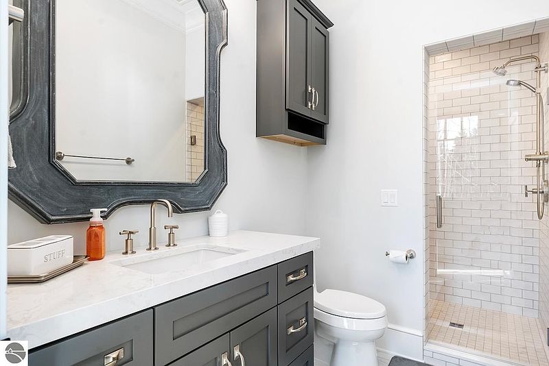 This is a well-lit bathroom featuring a gray vanity with a white countertop, a decorative mirror, and a glass-enclosed shower with white subway tiles. The color palette is neutral, creating a clean and modern aesthetic. The space appears functional and stylish, suitable for a guest or secondary bathroom.