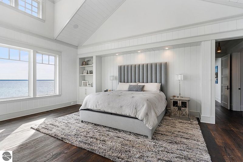 This primary bedroom features a serene and luxurious aesthetic with a large bed, a plush rug, and ample natural light from the windows overlooking the water. The room is decorated in neutral tones, with white walls and gray accents, creating a calming and inviting atmosphere. The built-in shelving and recessed lighting add to the room's sophisticated appeal.