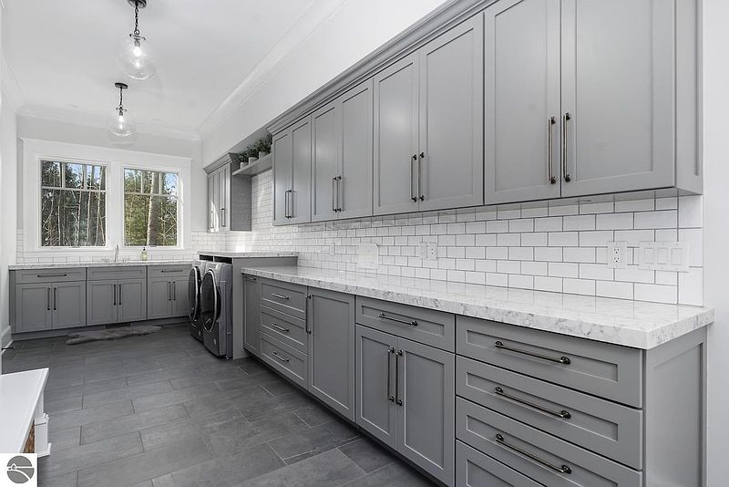 This is a well-organized laundry room featuring gray cabinetry with sleek hardware, a white subway tile backsplash, and a light-colored countertop. The room includes a washer and dryer set, along with ample storage space. The overall design is modern and functional, making it an appealing feature for potential buyers.