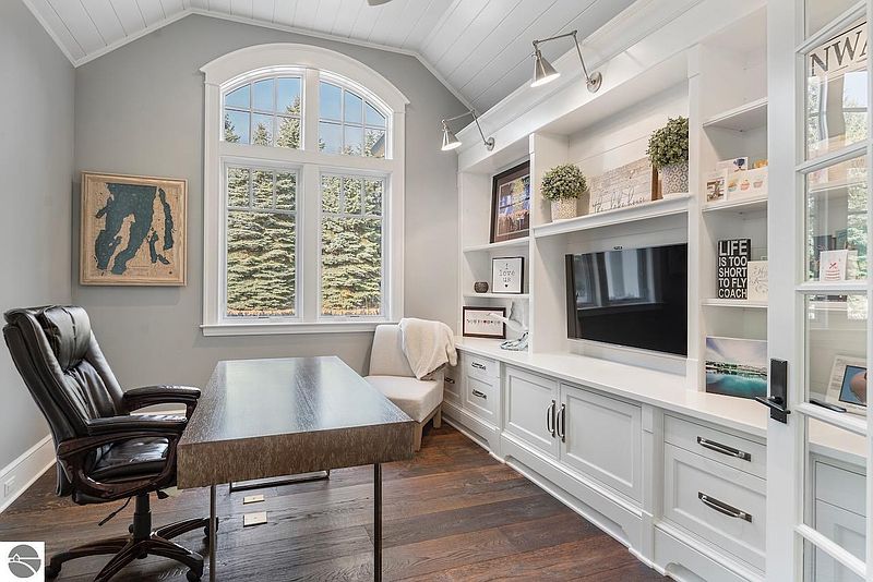 This is a well-appointed home office featuring a large desk, comfortable leather chair, and custom built-in shelving with a TV. The room has a bright and airy feel, thanks to the large window and light gray walls. The hardwood floors add warmth and sophistication to the space.