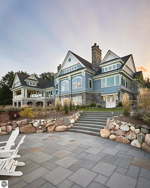 This image showcases the rear exterior of a luxurious home, featuring a stone patio, landscaped grounds, and a multi-level design with a combination of stone and siding. The property boasts multiple windows offering ample natural light and a grand staircase leading to the lawn, creating an inviting outdoor living space perfect for entertaining or relaxation.