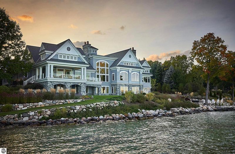 This image showcases the rear exterior of a luxurious waterfront home. The architectural style features a blend of traditional and modern elements, with large windows offering stunning views. The property includes a well-maintained lawn, stone landscaping, and direct access to the water, enhancing its appeal as a high-end real estate offering.