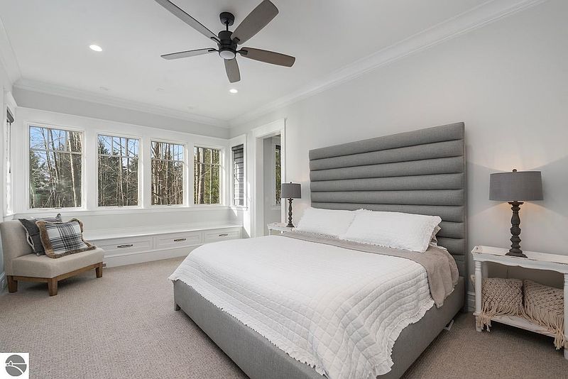 This is a bright and airy primary bedroom featuring a large bed with a gray upholstered headboard and white bedding. A bay window provides natural light and a view of the outdoors, while neutral tones create a calming atmosphere. The room is well-appointed with bedside tables and lamps.