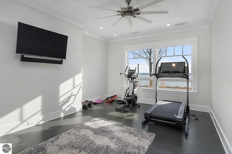 This home gym features a treadmill, elliptical machine, and free weights, providing a dedicated space for exercise. The room is well-lit with natural light from a large window offering a scenic view, and a mounted television provides entertainment during workouts. The dark flooring and neutral walls create a clean and functional environment.