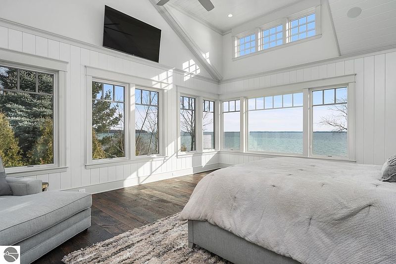 This is a bright and airy primary bedroom with a stunning waterfront view. The room features white paneled walls, large windows, and hardwood floors. A king-size bed with a light gray comforter is the focal point, complemented by a cozy chaise lounge. A large television is mounted on the wall.