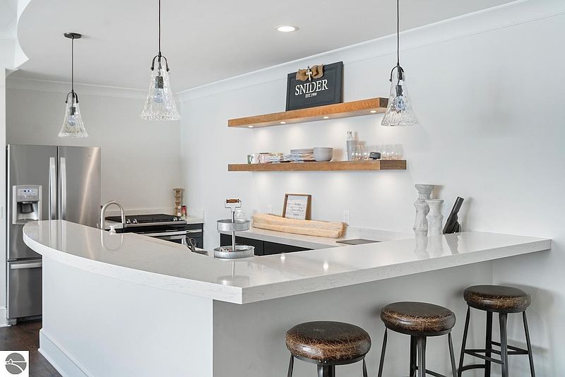 This kitchen features a curved countertop with bar seating, stainless steel appliances, and open shelving. The design incorporates a mix of white and dark cabinetry, complemented by pendant lighting and decorative elements. The space appears modern and well-lit, creating an inviting atmosphere.