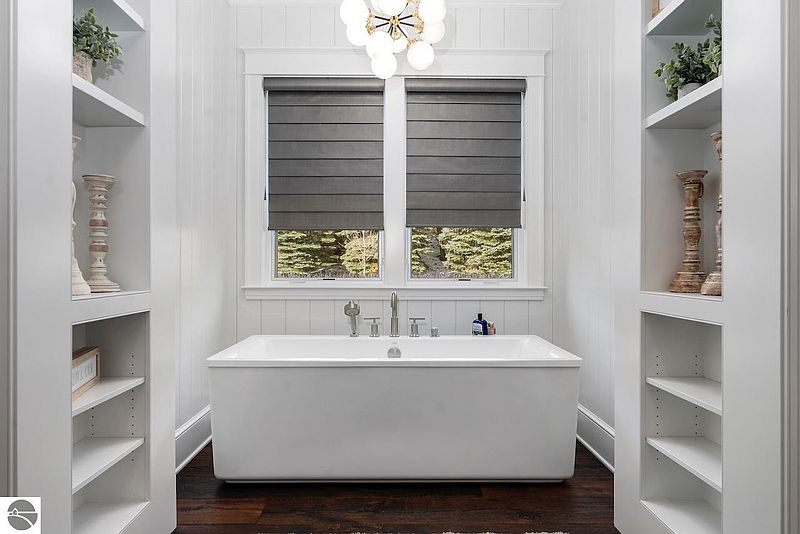 This is a luxurious primary bathroom featuring a freestanding white bathtub centered between two built-in shelving units. Natural light filters through the window with gray roman shades, illuminating the white paneled walls and dark wood flooring. The overall impression is clean, modern, and spa-like.