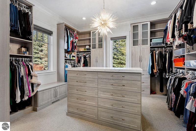 This is a spacious walk-in closet featuring custom shelving and drawers, providing ample storage for clothing and accessories. A central island with a countertop offers a convenient space for folding or organizing items. The closet is well-lit with a modern chandelier, and the overall design is elegant and functional.