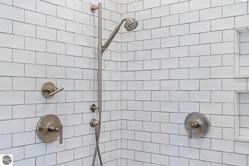 This image showcases a modern primary bathroom shower with white subway tile and brushed nickel fixtures. The shower features a handheld shower head and sleek, minimalist controls. The overall impression is clean, bright, and luxurious.