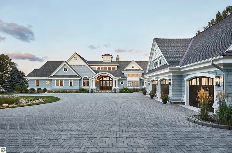 This is a front view of a large, luxurious home with a sprawling driveway. The house features a combination of light blue siding and gray shingles, with multiple gables and a prominent entryway. A three-car garage is attached to the side, and the landscaping is well-maintained, creating an inviting curb appeal.