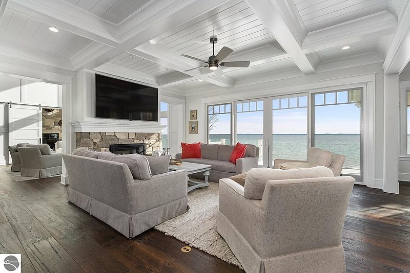 This is a bright and airy living room with a view of the water. The room features a stone fireplace with a mounted television, comfortable seating, and hardwood floors. The coffered ceiling and large windows add to the luxurious feel of the space.