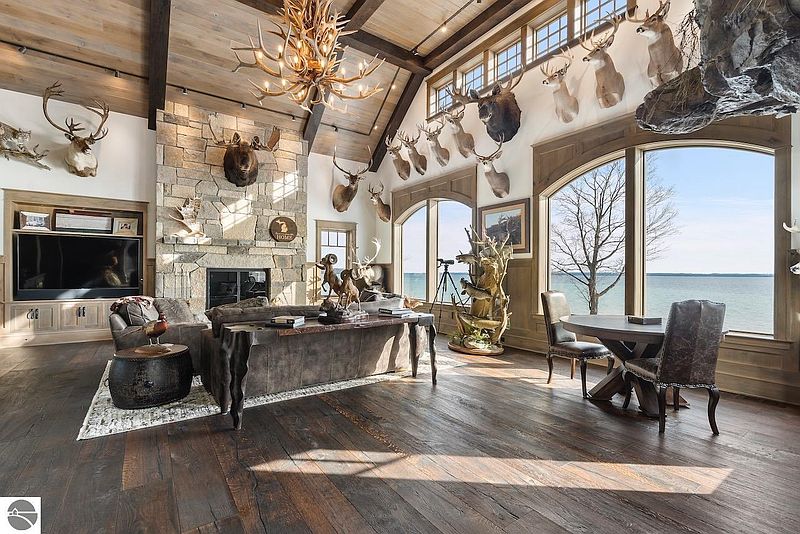 This grand living room features a high vaulted ceiling with exposed beams, a stone fireplace, and large windows offering a scenic water view. The room is decorated with numerous taxidermied animals, creating a rustic and unique atmosphere. Hardwood floors and a large chandelier add to the room's luxurious feel.