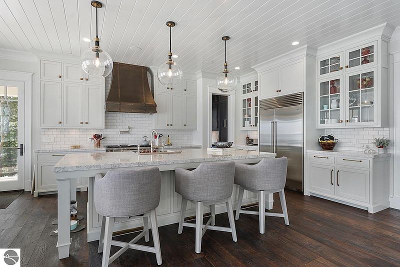 This is a bright and modern kitchen featuring white cabinetry, a large island with seating, and stainless steel appliances. The kitchen is well-lit with pendant lighting and natural light, creating a welcoming and functional space. Dark hardwood floors add a touch of elegance and contrast to the light color palette.