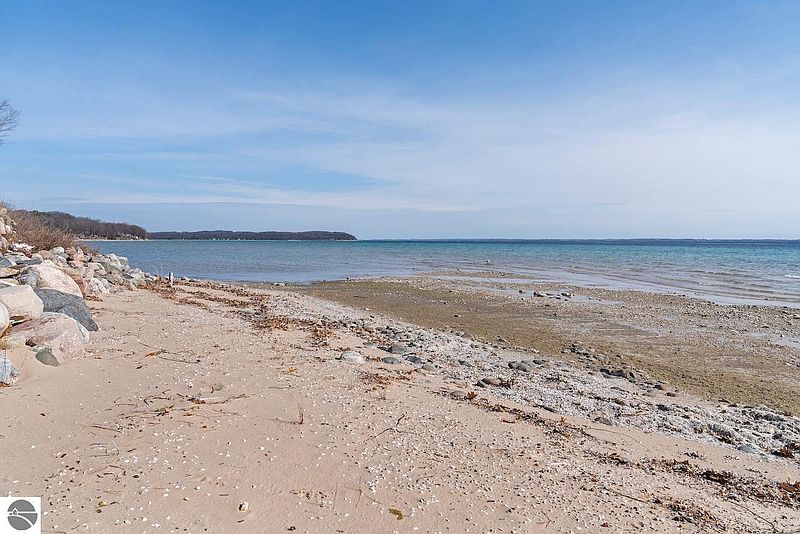 This image showcases a serene beach scene with a sandy shoreline meeting calm, turquoise water under a clear blue sky. Large rocks line the edge of the beach, transitioning into a sandy area with scattered shells and pebbles. The scene evokes a sense of tranquility and natural beauty, highlighting the property's access to a waterfront environment.