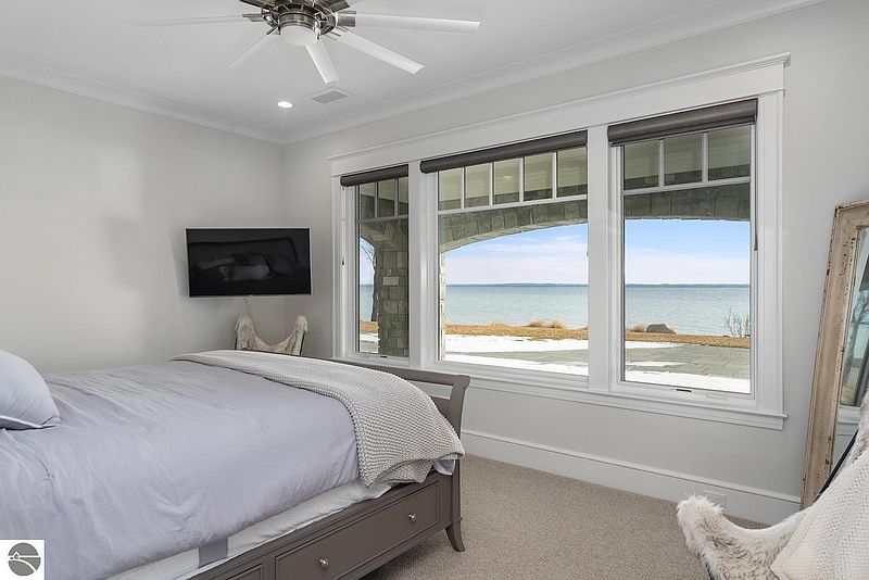 This bedroom features a serene view of the water through a large window, complemented by a neutral color palette and a comfortable bed. A wall-mounted television adds a modern touch, while the overall design creates a relaxing and inviting atmosphere. The room is well-lit and appears spacious.
