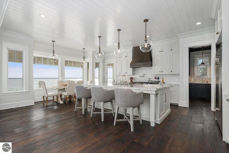 This is a bright and spacious kitchen featuring a large island with bar seating, white cabinetry, and pendant lighting. The kitchen opens to a dining area with large windows offering a scenic water view. Dark hardwood floors add warmth and contrast to the light color palette, creating an inviting and luxurious atmosphere.