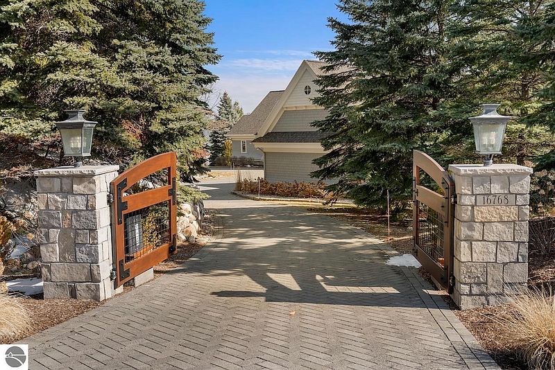 This image showcases an inviting entryway to a property, featuring a brick-paved driveway leading through an open wooden gate flanked by stone pillars with elegant lanterns. The house is partially visible in the background, suggesting a well-maintained and private estate. Mature trees frame the entrance, adding to the property's curb appeal and sense of seclusion.