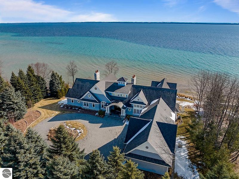 This aerial view showcases a luxurious waterfront home with a sprawling layout and a dark roof. The property features a circular driveway, mature trees, and direct access to a beautiful body of water with varying shades of blue. The home's architecture is impressive, suggesting high-end finishes and ample living space.