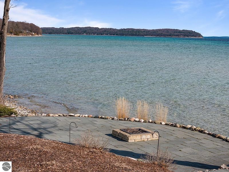 This image showcases a serene patio area overlooking a tranquil body of water. The patio is constructed with large, gray pavers and features a stone fire pit, creating an inviting outdoor living space. The scene evokes a sense of relaxation and connection with nature, making it an appealing feature for potential buyers.