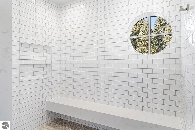This is a bright and modern primary bathroom featuring white subway tile walls, a built-in bench, and a unique circular window providing natural light and a view of the trees outside. The clean lines and minimalist design create a spa-like atmosphere, perfect for relaxation.