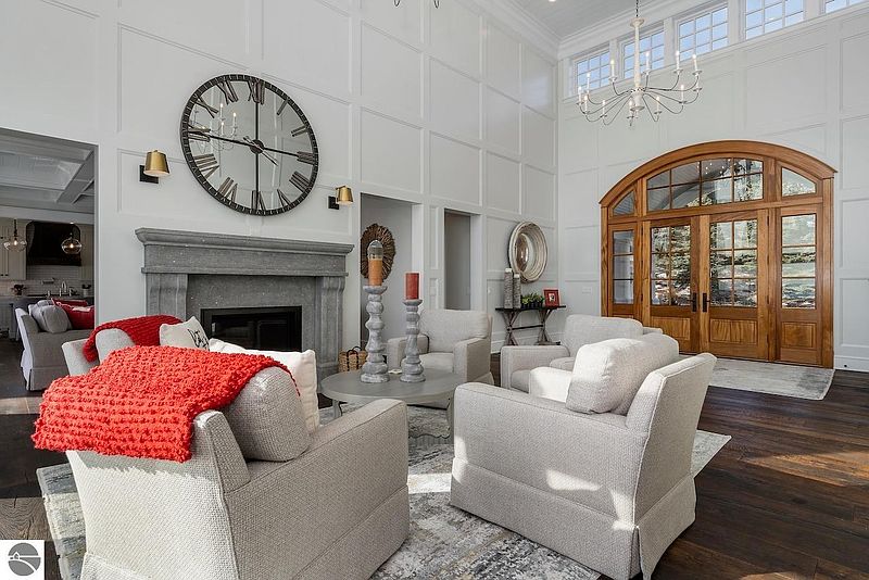 This is a bright and spacious living room featuring a high ceiling, a large fireplace with a stone mantel, and elegant wood flooring. The room is furnished with comfortable armchairs and a round coffee table, creating an inviting and sophisticated atmosphere. A large clock above the fireplace serves as a focal point.