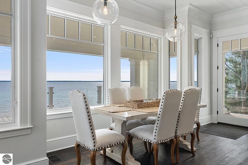 This is an interior shot of a dining room featuring a white dining table with upholstered chairs. Large windows offer a scenic view of the water, enhancing the room's bright and airy atmosphere. The room is decorated in a neutral color palette, creating a sophisticated and inviting space.