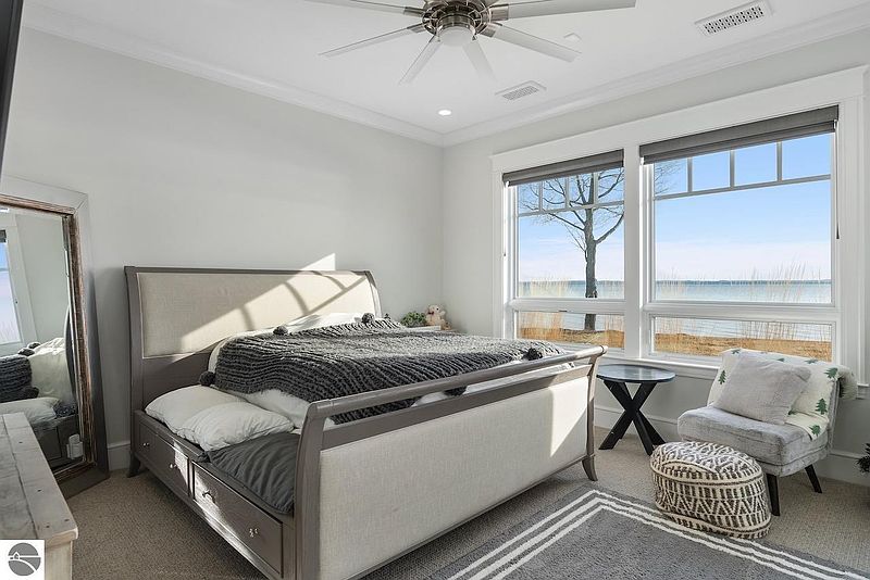 This bedroom features a neutral color palette with a gray bed frame and headboard, complemented by a gray and white rug. Large windows offer a view of the water, enhancing the room's serene atmosphere. The space is well-lit and styled with comfortable seating, creating a relaxing and inviting environment.
