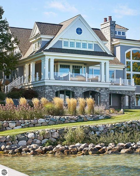This is a stunning front view of a luxurious home with a stone foundation, gray siding, and a prominent gable roof. The property features a spacious balcony with glass railings, manicured landscaping, and a stone retaining wall leading down to the water. The architectural details and serene waterfront setting create an impressive and desirable residence.