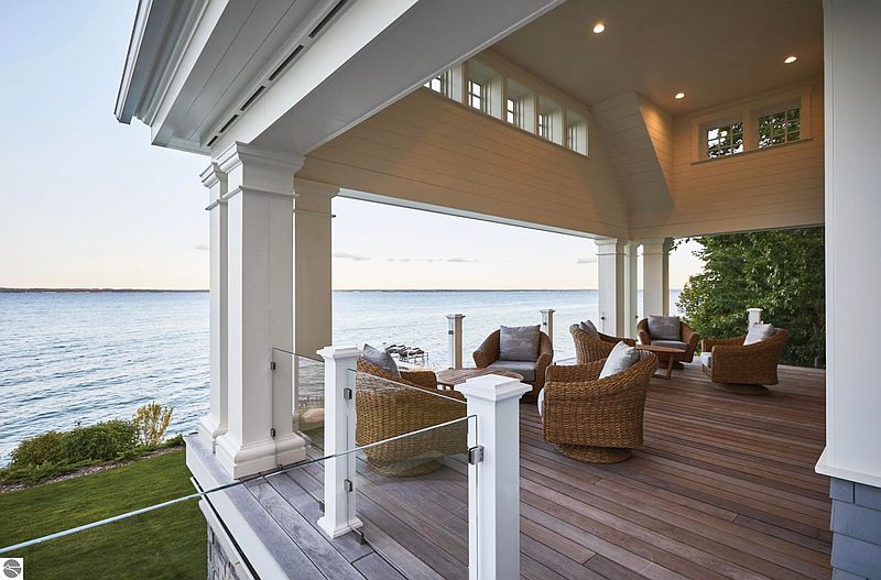 This image showcases a luxurious outdoor patio or deck area with stunning waterfront views. The space is furnished with comfortable wicker seating and features a wooden deck, glass railings, and substantial white support columns. The design emphasizes relaxation and enjoying the scenic surroundings, making it an ideal selling point for a property.