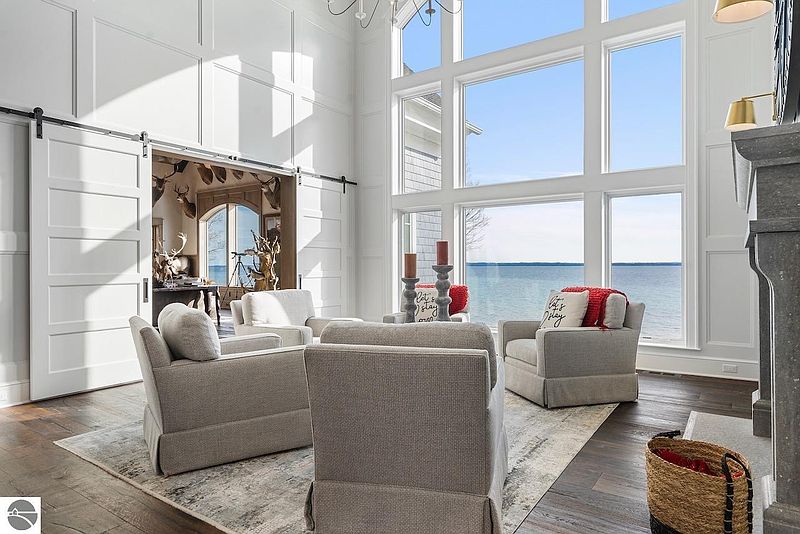 This is a bright and airy living room with a stunning view of the water through large windows. The room features comfortable seating, a neutral color palette, and elegant architectural details such as paneled walls and a sliding barn door. The space exudes a sense of luxury and tranquility, perfect for relaxation and entertaining.