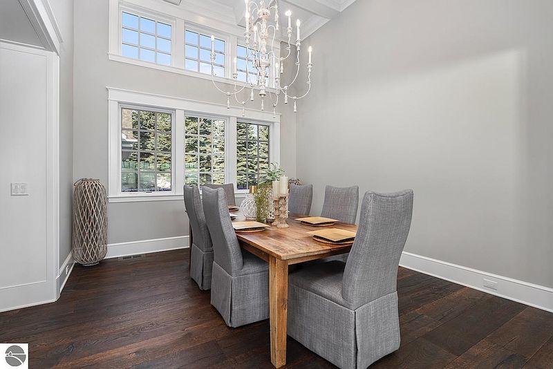 This is a well-lit dining room featuring a wooden table with six upholstered chairs. The room has a neutral color palette with gray walls and dark hardwood floors. A decorative chandelier hangs above the table, and large windows provide natural light, creating a warm and inviting atmosphere.
