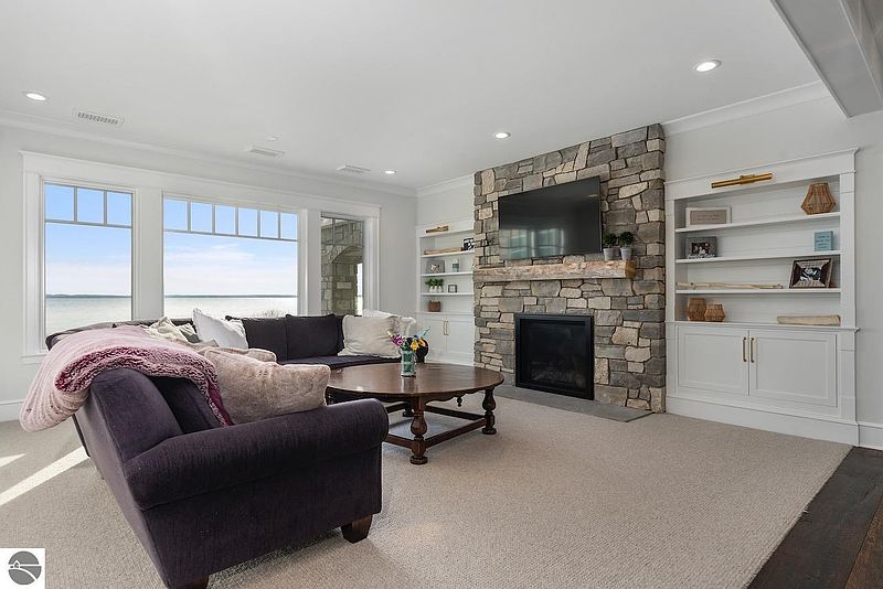 This living room features a stone fireplace with a mounted television, built-in shelving units, and a comfortable sectional sofa. Large windows offer a view of the water, creating a bright and inviting atmosphere. The neutral color palette and plush carpeting contribute to a cozy and elegant feel.